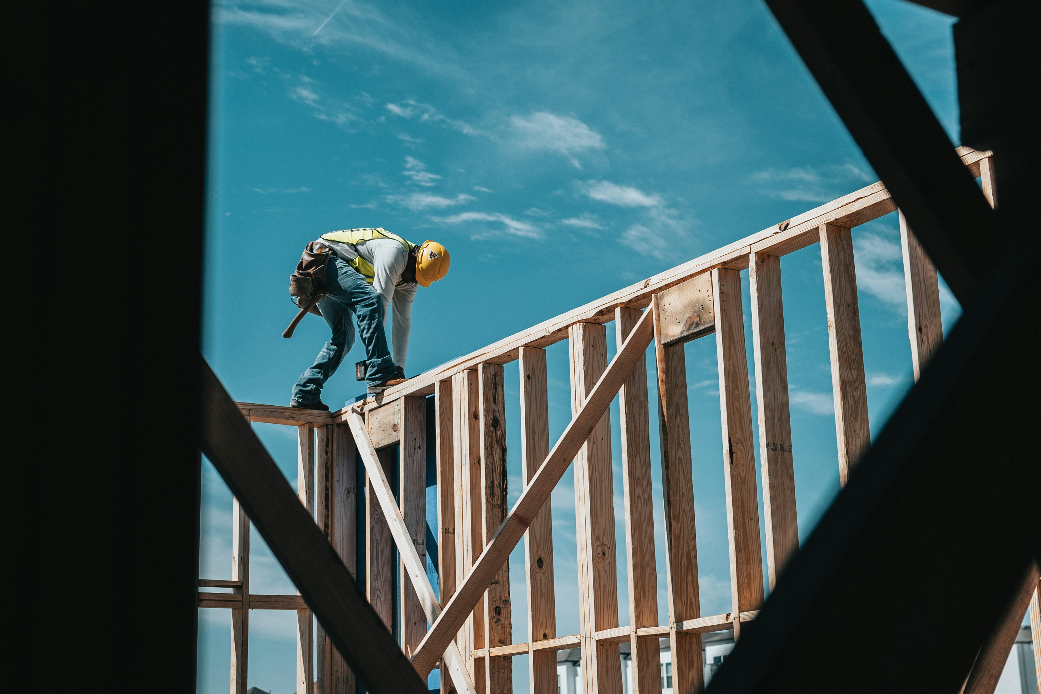 A worker in a high-vis jacket standing on top of a new house frame, nailing it together, against a bright background.
